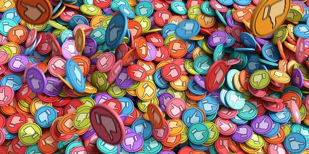 An abstract image of lots of pastel toned multi-coloured discs with 'Like' and 'Dislike' thumbs up and thumbs down icons on them. Some discs are in mid air with motion blur and are falling onto a large pile of discs. Concepts: social media issues.